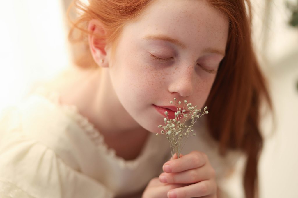 Niña pelirroja oliendo una pequeña flor con los ojos cerrados, en luz natural.